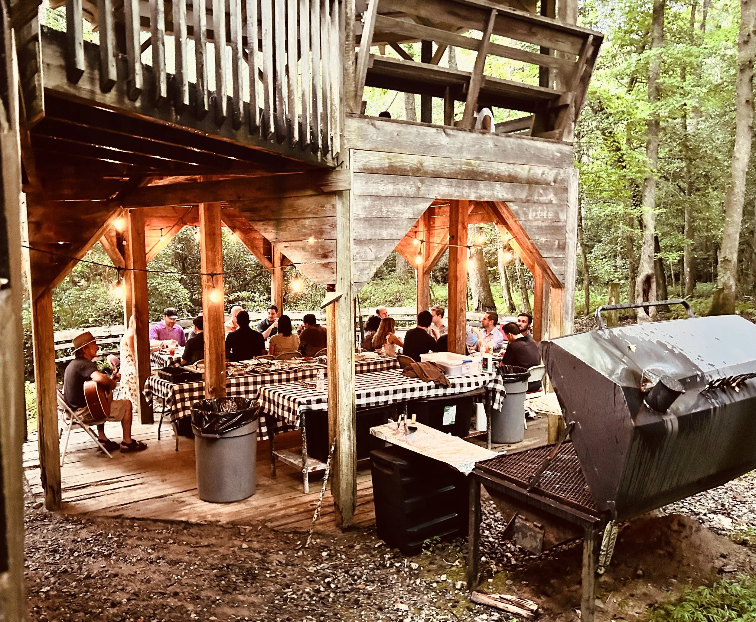Outdoor gathering under a wooden treehouse with string lights, people sitting at tables, some playing guitar, surrounded by trees.
