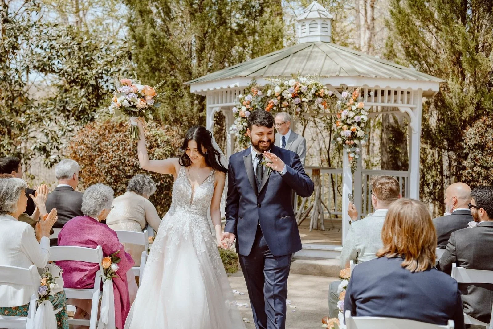 A newlywed couple walking down the aisle at their outdoor wedding ceremony, surrounded by seated guests, with a decorated gazebo in the background, during daytime.