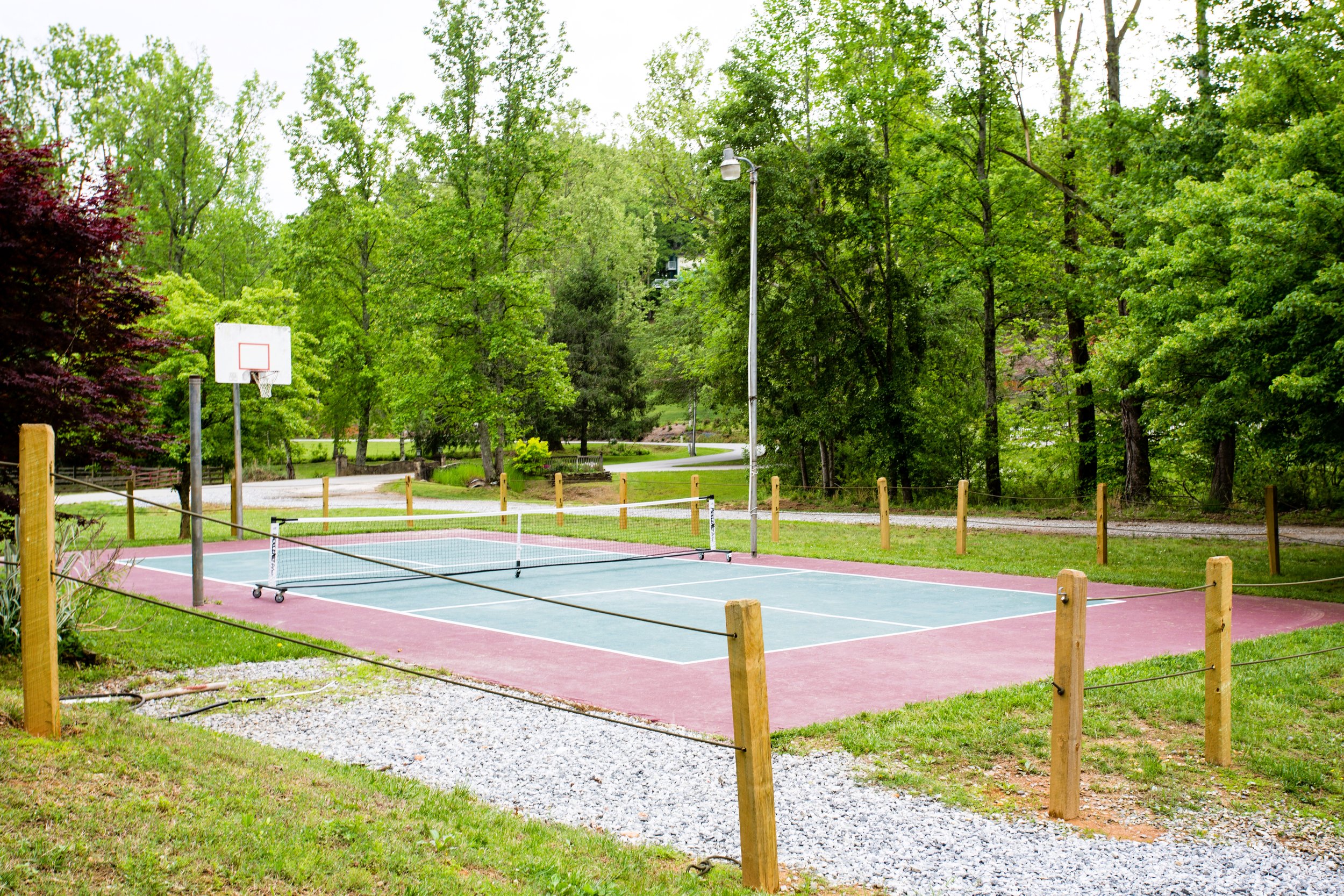 A small outdoor volleyball court with a net, surrounded by a wooden fence, nearby is a basketball hoop, and tall green trees in the background.