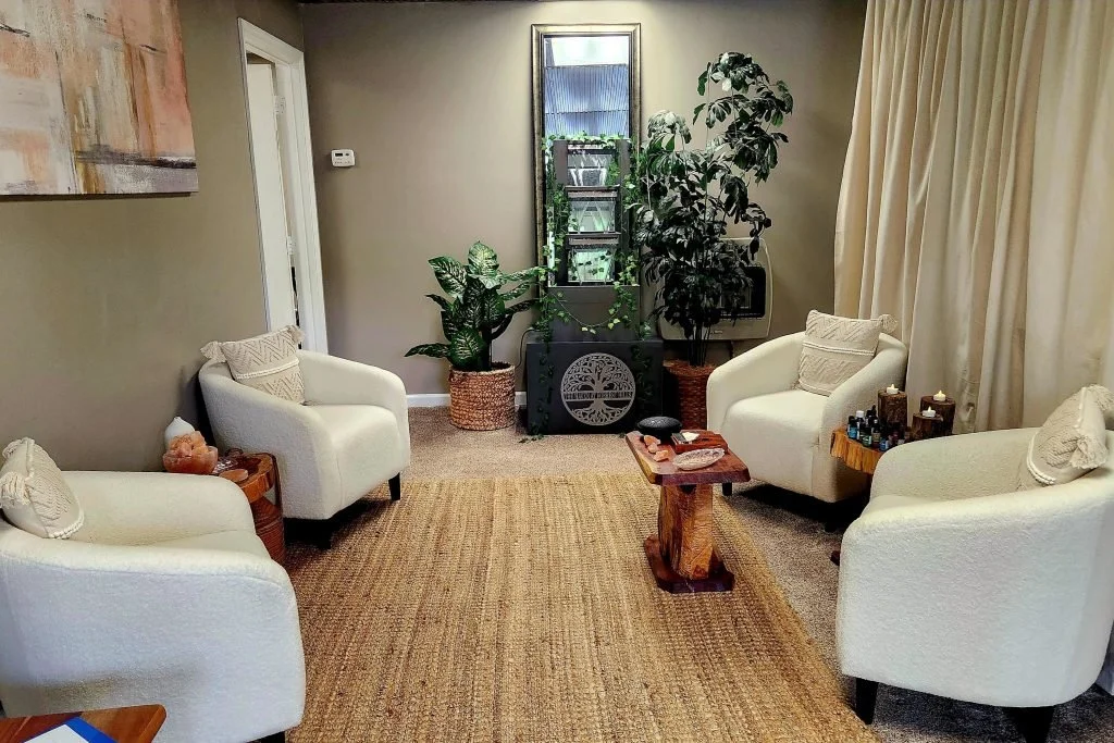 A cozy living room with four white armchairs arranged around a central wooden coffee table, a woven rug on the floor, large green plants in baskets near a window, and a shelf with plants, books, and decorative items against a gray wall.