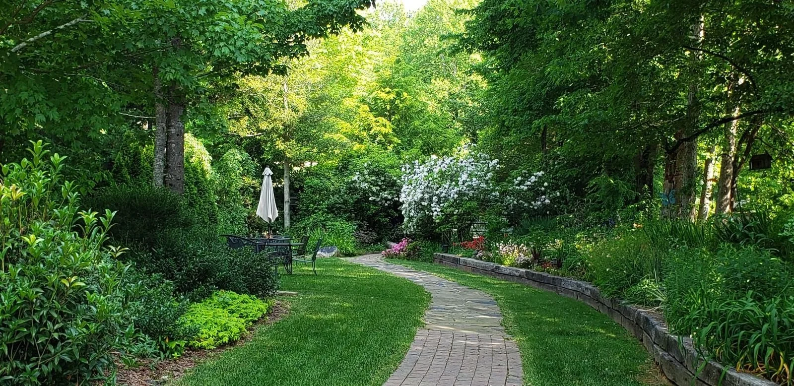 A lush green garden with a winding stone and brick pathway, surrounded by flowering bushes, trees, and outdoor furniture including a white patio umbrella.
