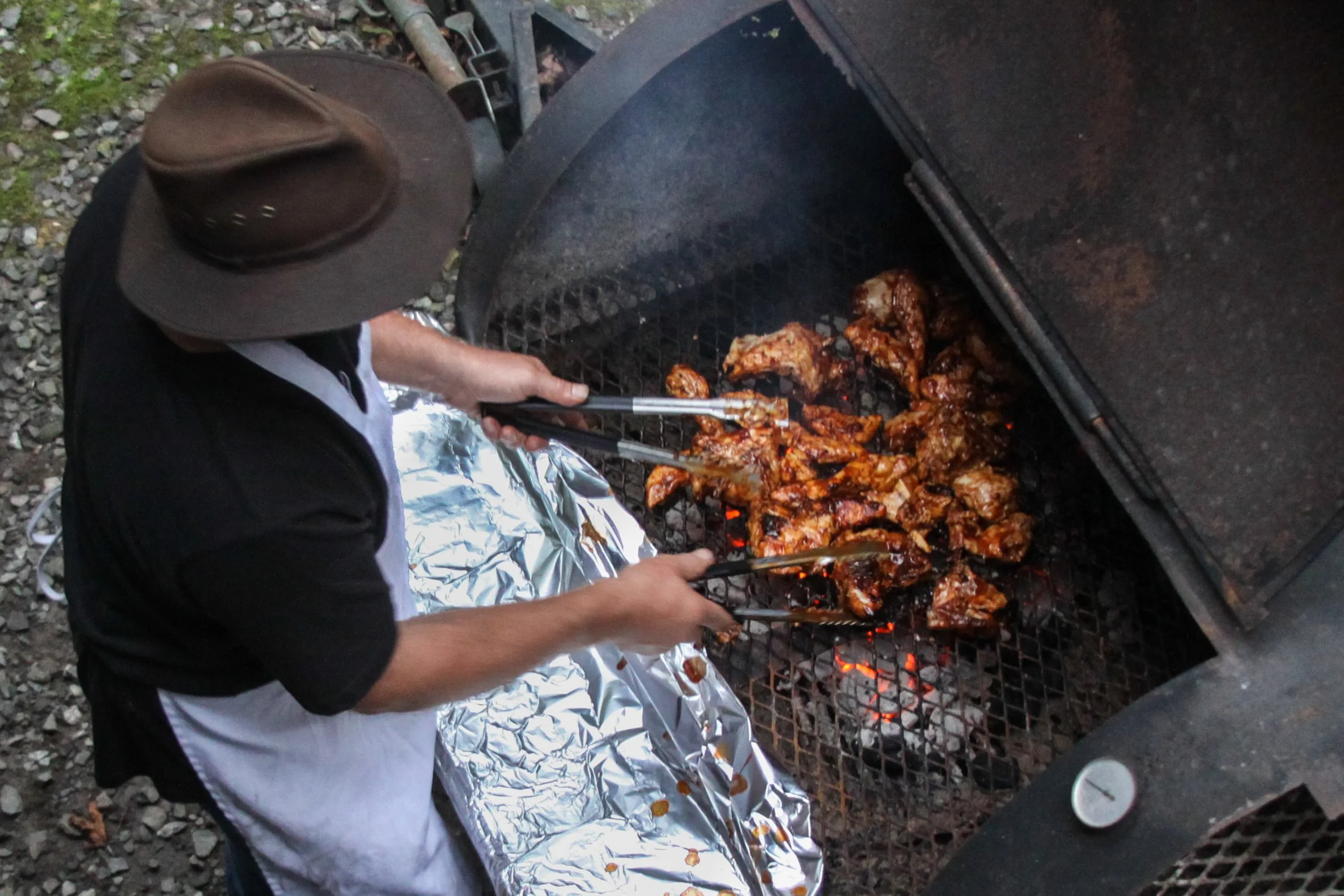 Person wearing a cowboy hat and apron grilling marinated chicken pieces on a barbecue grill outdoors.
