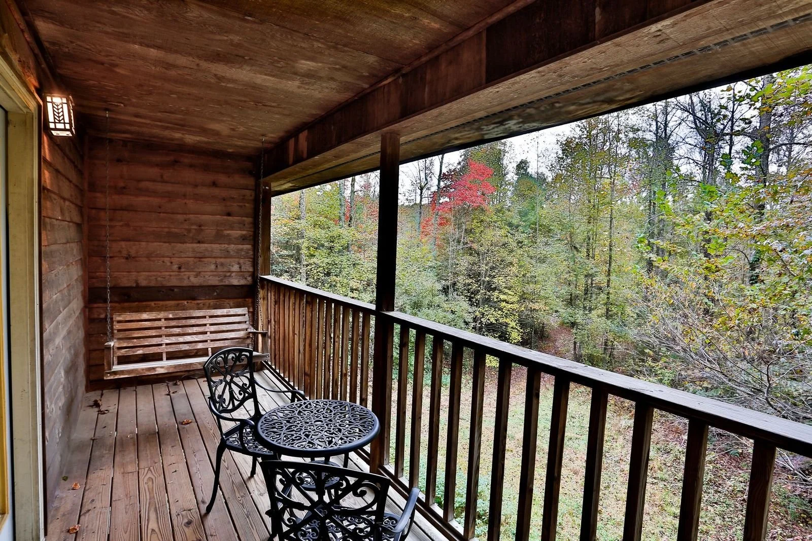 Wooden balcony with a wrought iron table and chairs, a wooden bench swing, overlooking a forest with trees showing fall colors.