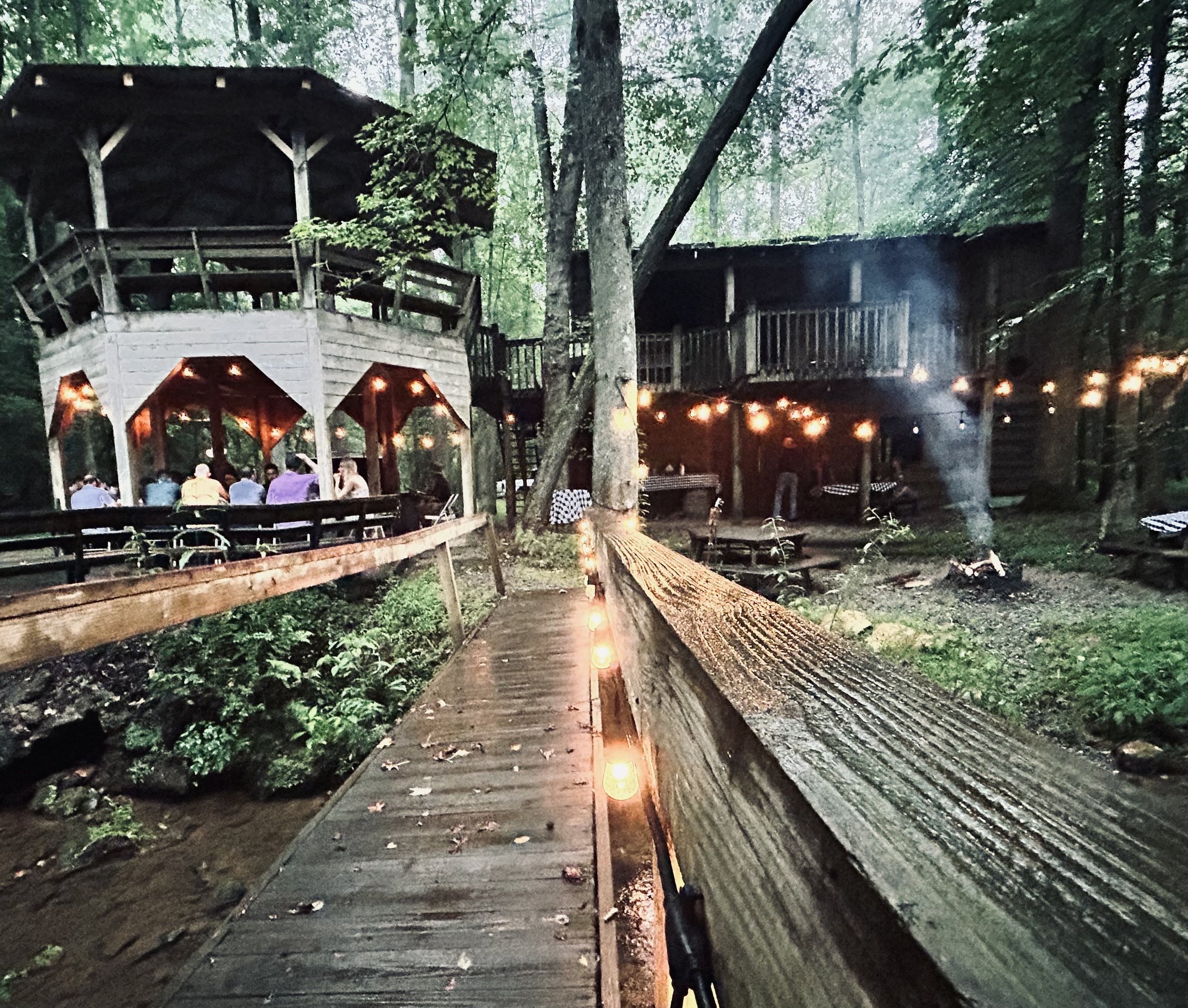 A treehouse built among trees in a forest, illuminated by string lights, with people sitting and standing on the deck below.