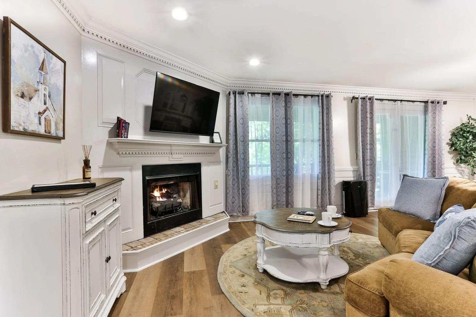 Living room with fireplace, wall-mounted TV, beige and gray couches, wooden coffee table with cups and notebooks, curtains, potted plant, and hardwood flooring.