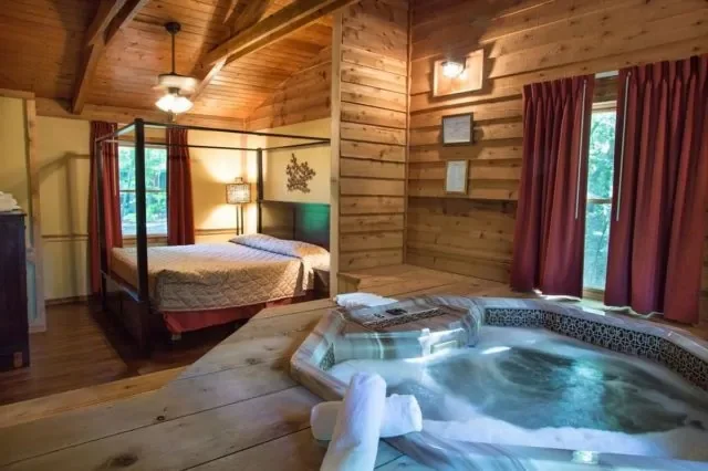 A cozy wooden cabin bedroom with a four-poster bed and a hot tub in the corner, along with red curtains and framed pictures on the wall.