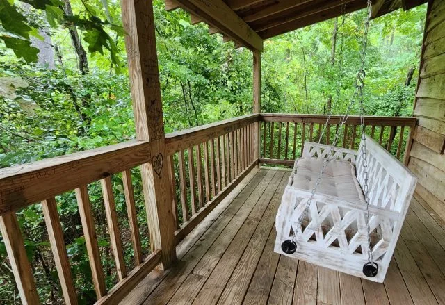 A wooden porch with a railing, overlooking a green forest, with a white wooden porch swing hanging from the ceiling.