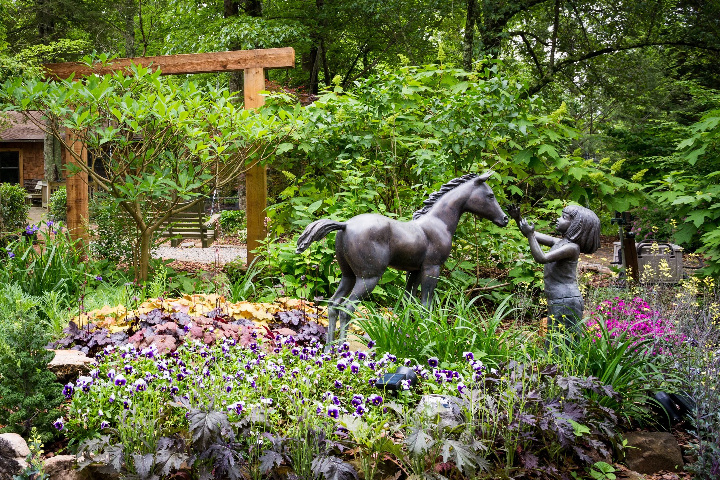 A garden scene with a metal sculpture of a girl and a horse, surrounded by lush green foliage and colorful flowers.
