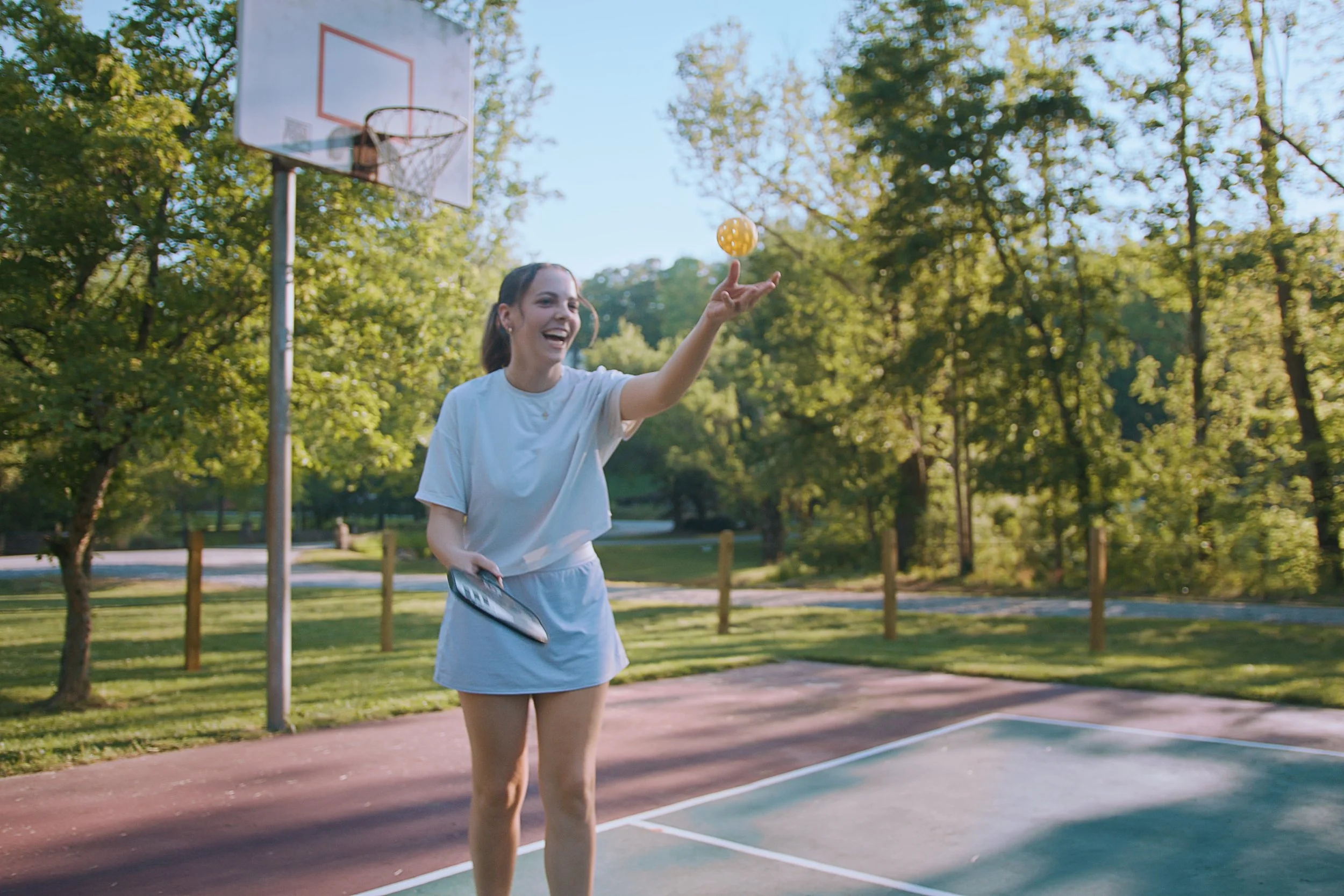 A woman on an outdoor basketball court hitting a yellow ball with her finger. She is smiling, wearing a light-colored t-shirt and shorts, with a basketball hoop in the background and trees surrounding the court.