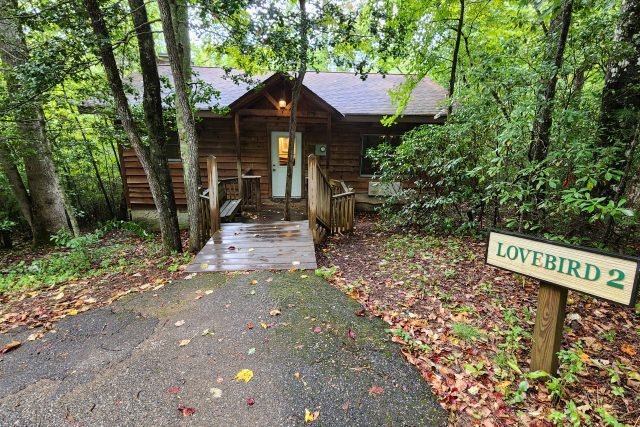 A wooden cabin in a wooded area with a ramp leading to the door, and a sign that reads 'LOVE BIRD 2' to the right.