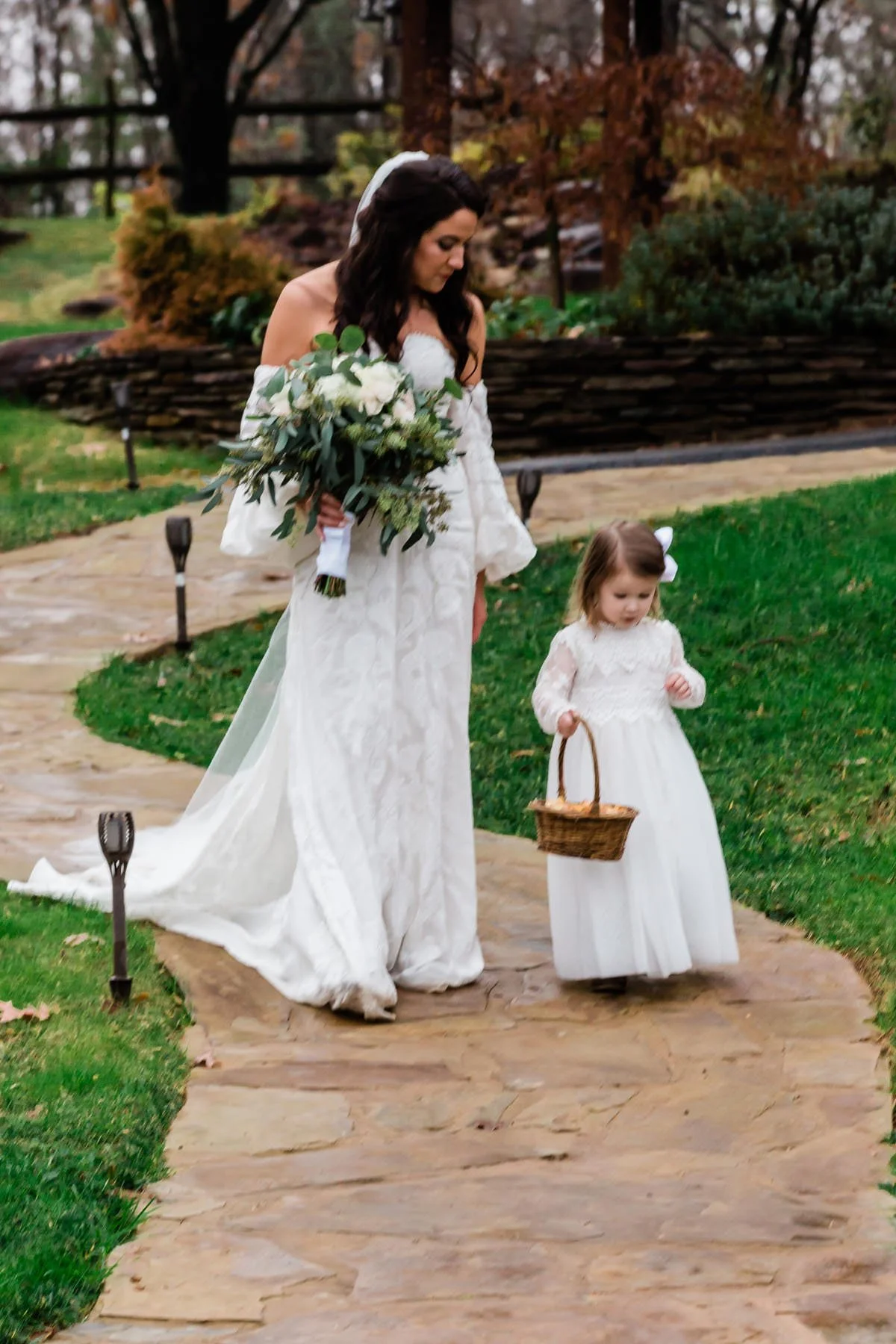 A woman in a wedding dress holding a bouquet and a young girl in a white dress carrying a small basket walking on a stone pathway in a garden.
