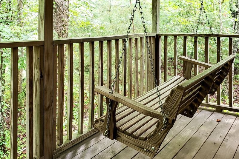 Wooden porch with a swing and railing overlooking a wooded area.