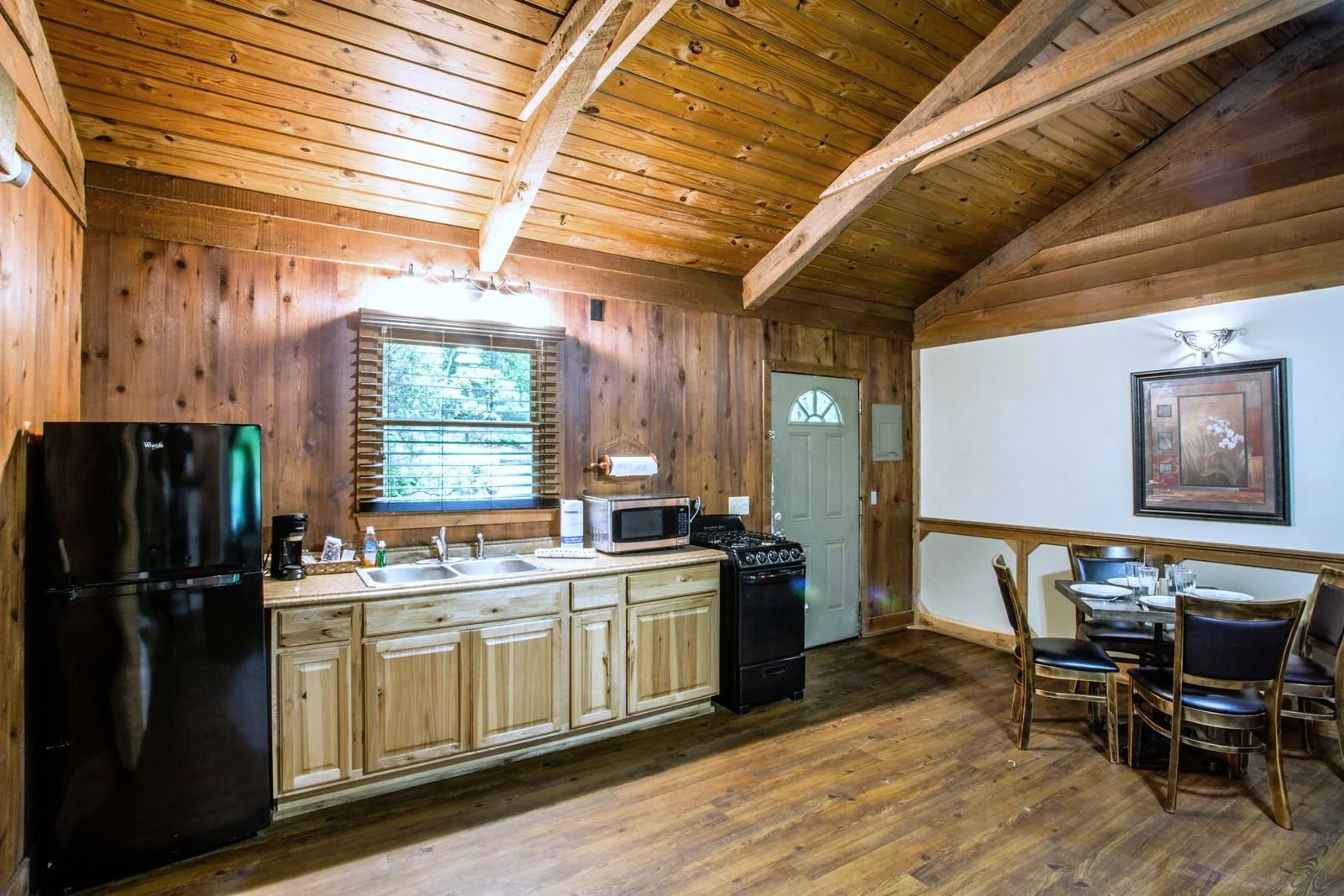 A rustic kitchen and dining area with wooden walls and ceiling, featuring a black refrigerator, microwave, stove, and a round dining table with chairs.