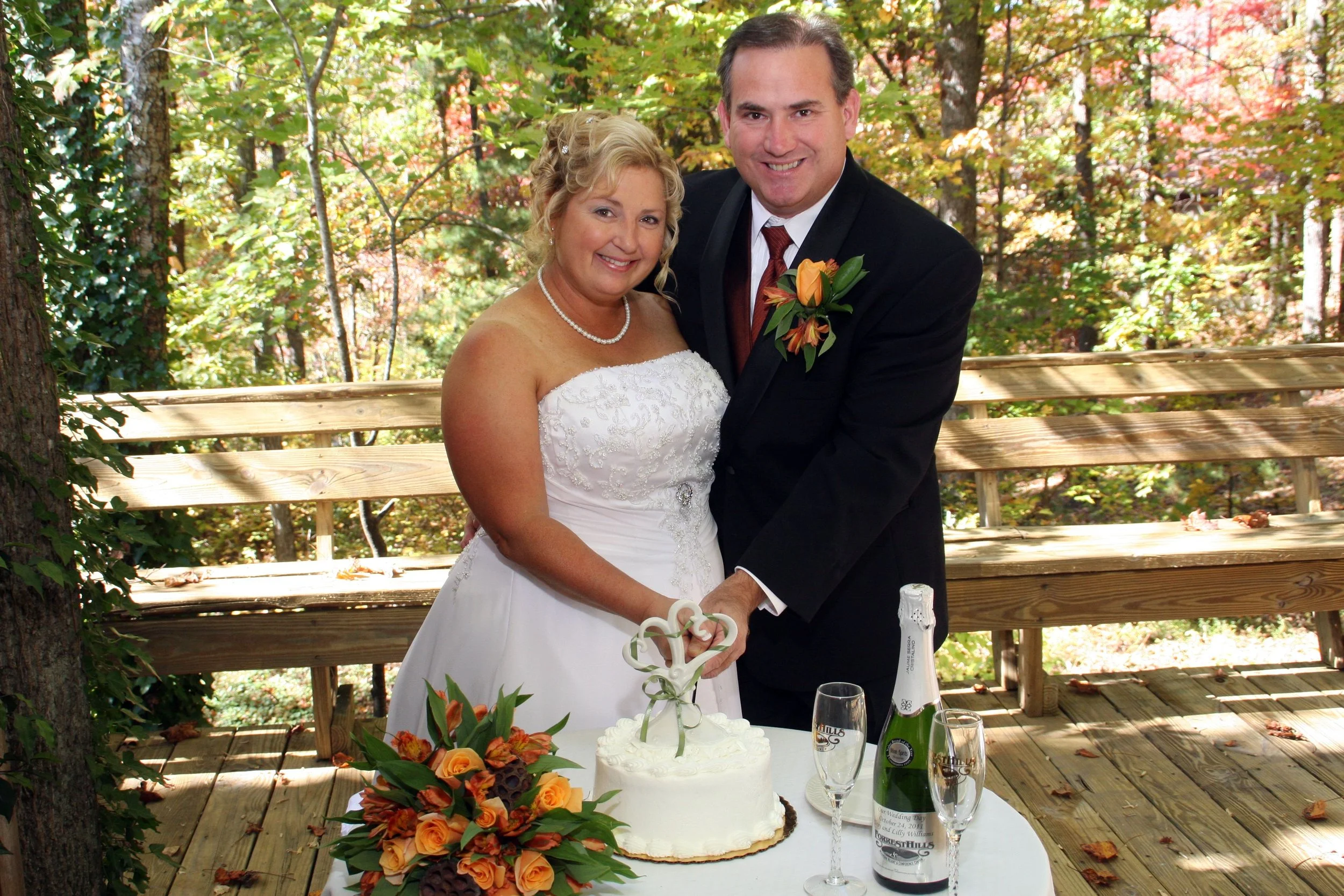 A bride and groom stand together outside on a wooden deck, celebrating their wedding with a cake and drinks on a table. The bride wears a white wedding dress and pearl necklace, and the groom is in a black suit with a red tie and boutonniere. The bac