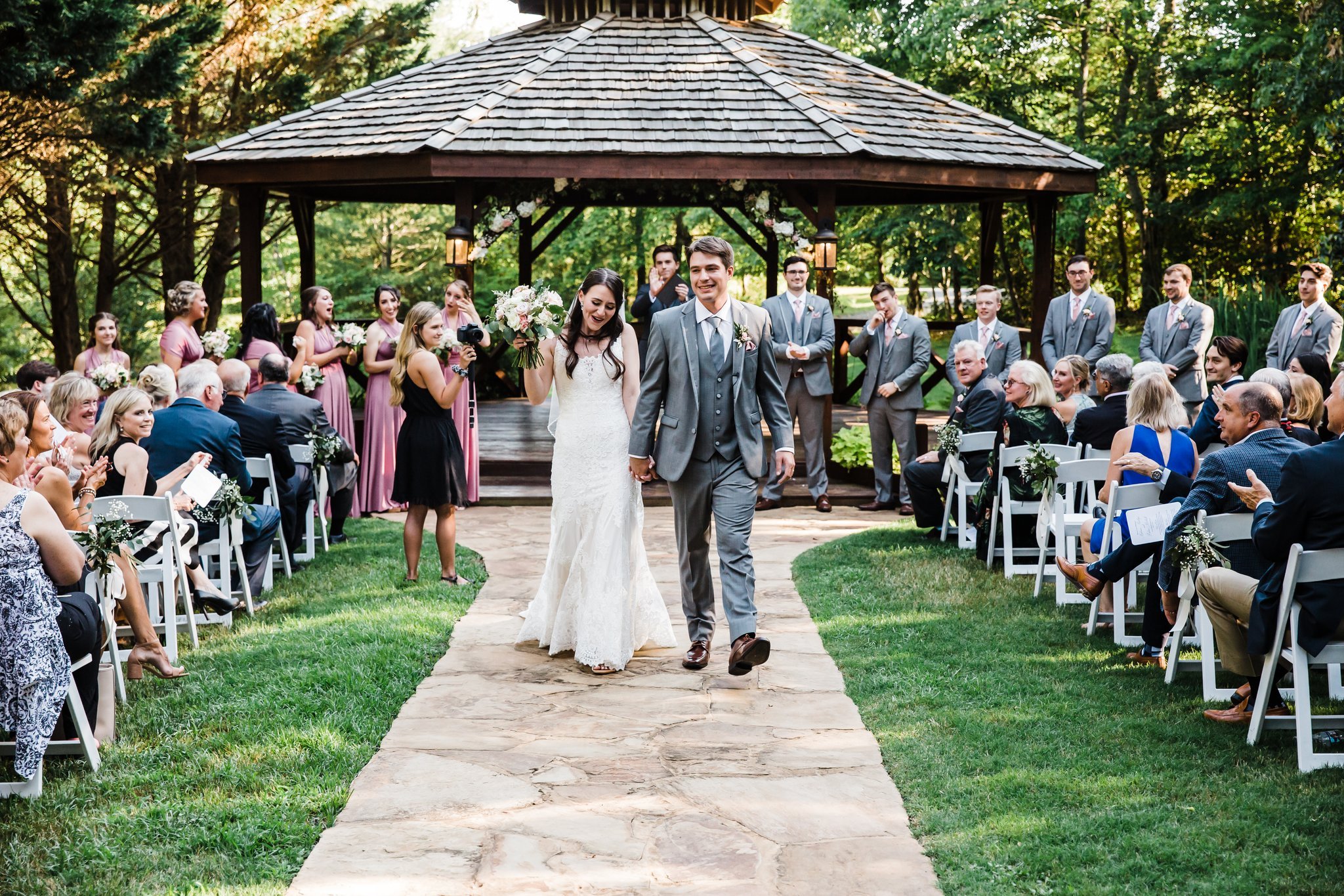 A bride and groom walking down the aisle during a wedding ceremony in an outdoor garden setting, with guests seated on either side and a gazebo in the background.