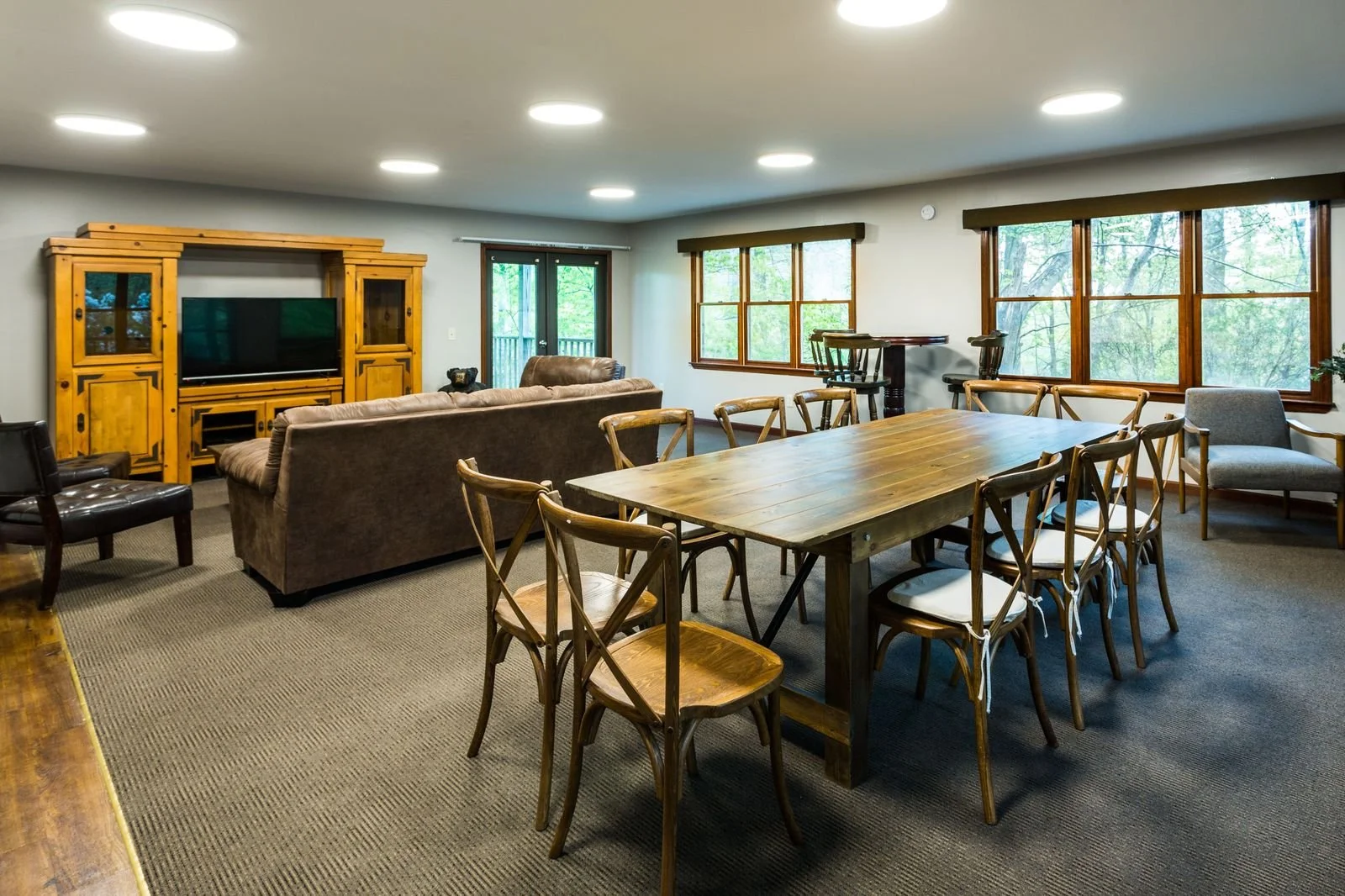 Living and dining room with a large wooden table and chairs, a brown couch, a TV in a wooden cabinet, and large windows overlooking trees.