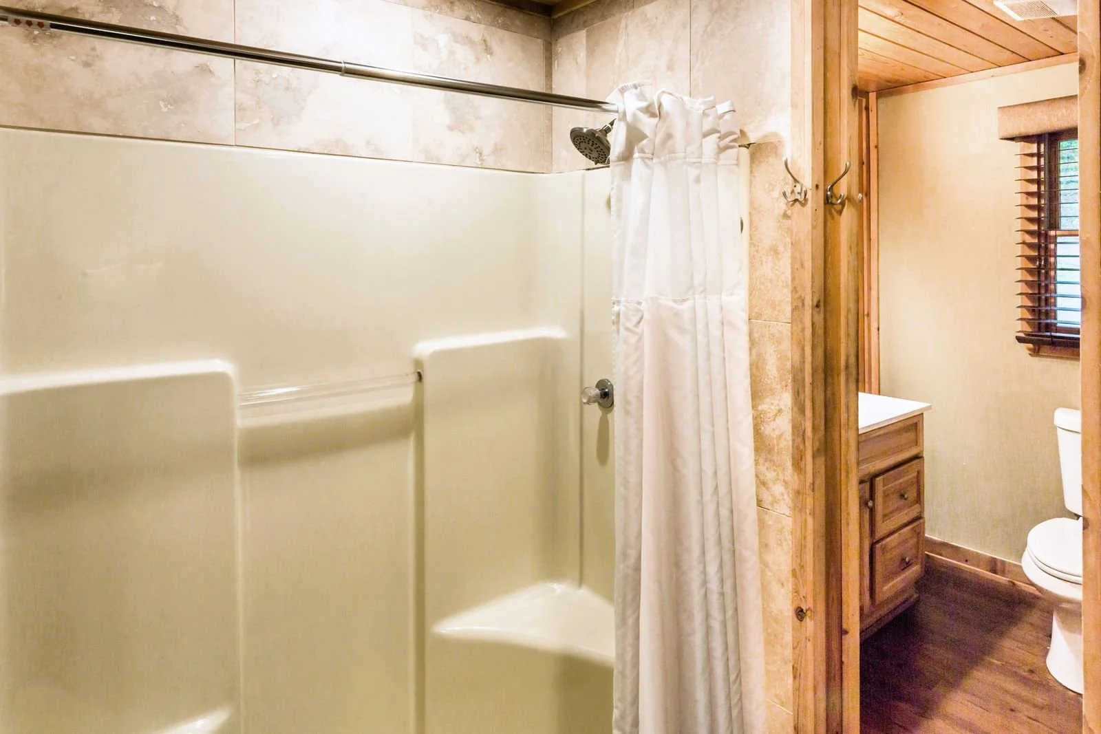 Bathroom with a walk-in shower with a cream-colored folding door, white shower curtain, beige tile walls, a wooden vanity, a window with blinds, and a toilet.