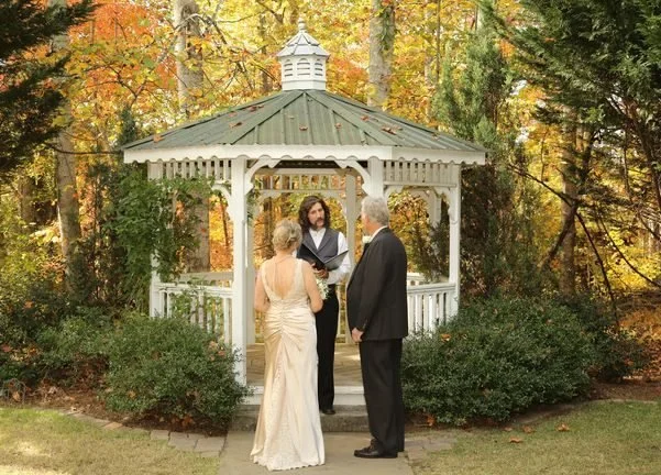 A wedding ceremony taking place outdoors near a white gazebo with autumn trees in the background. The bride and groom stand facing each other, with a officiant reading from a book.
