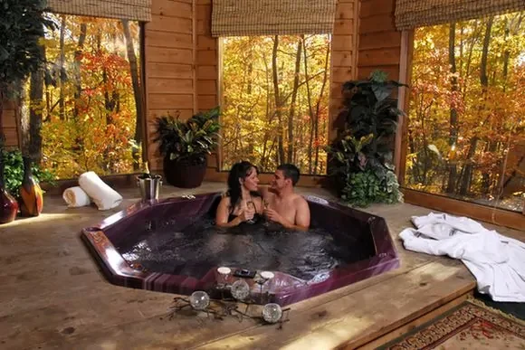 A couple sitting in a hot tub inside a wooden cabin with large windows showing autumn trees outside.