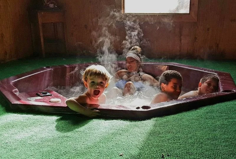 Four children and an adult sitting in a bubbling hot tub inside a wooden room, with steam rising from the water.
