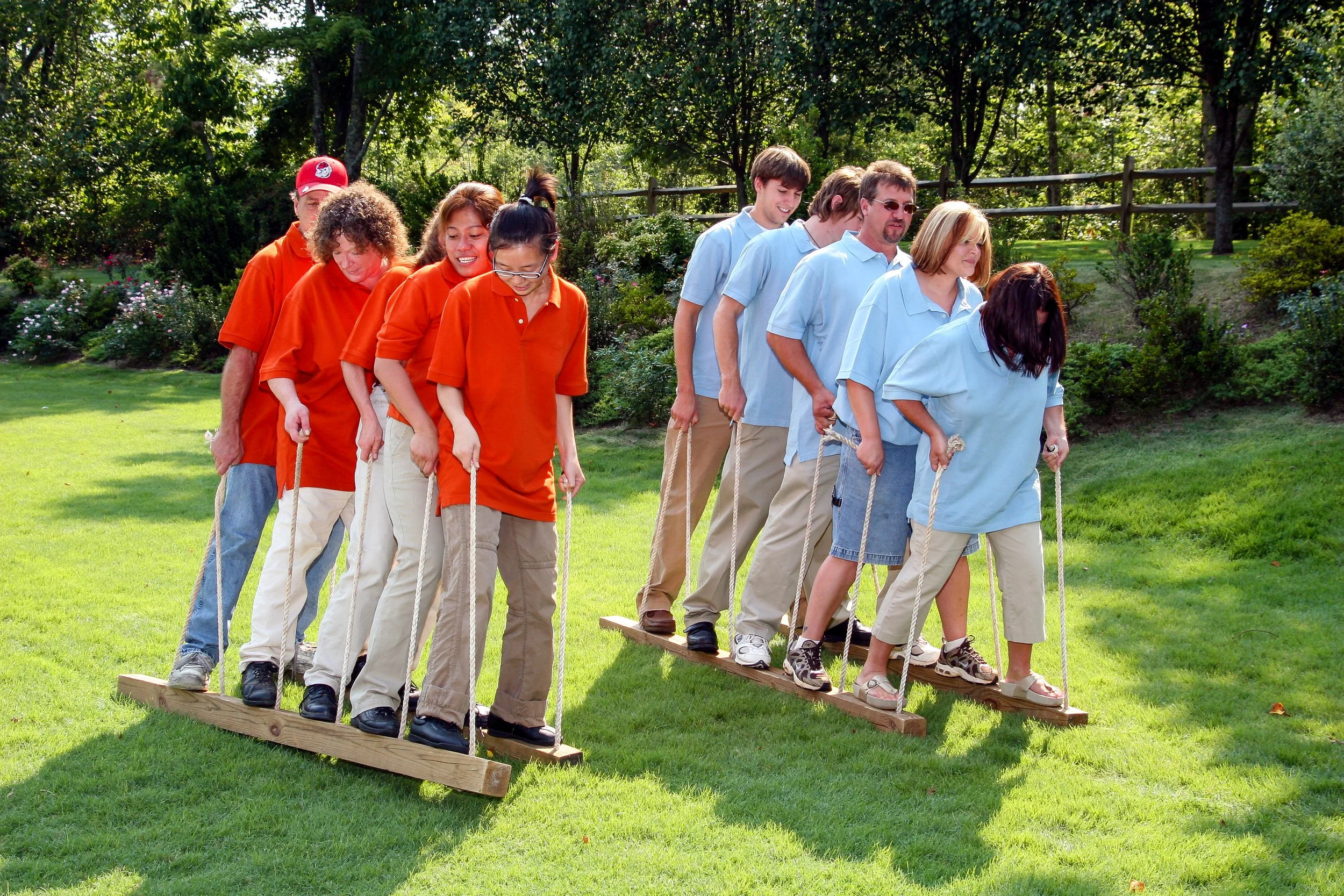 A group of people participating in a team-building outdoor activity, walking on wooden planks while holding ropes for balance in a park.