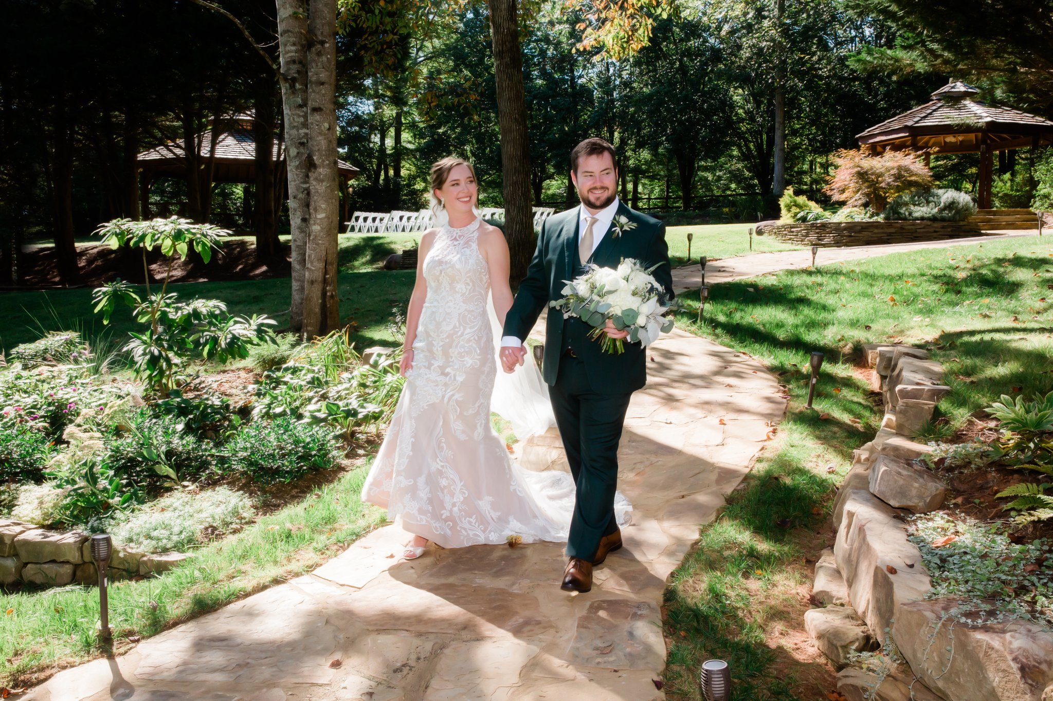 A bride and groom walking hand in hand outdoors during a wedding ceremony in a garden setting with trees and potted plants.