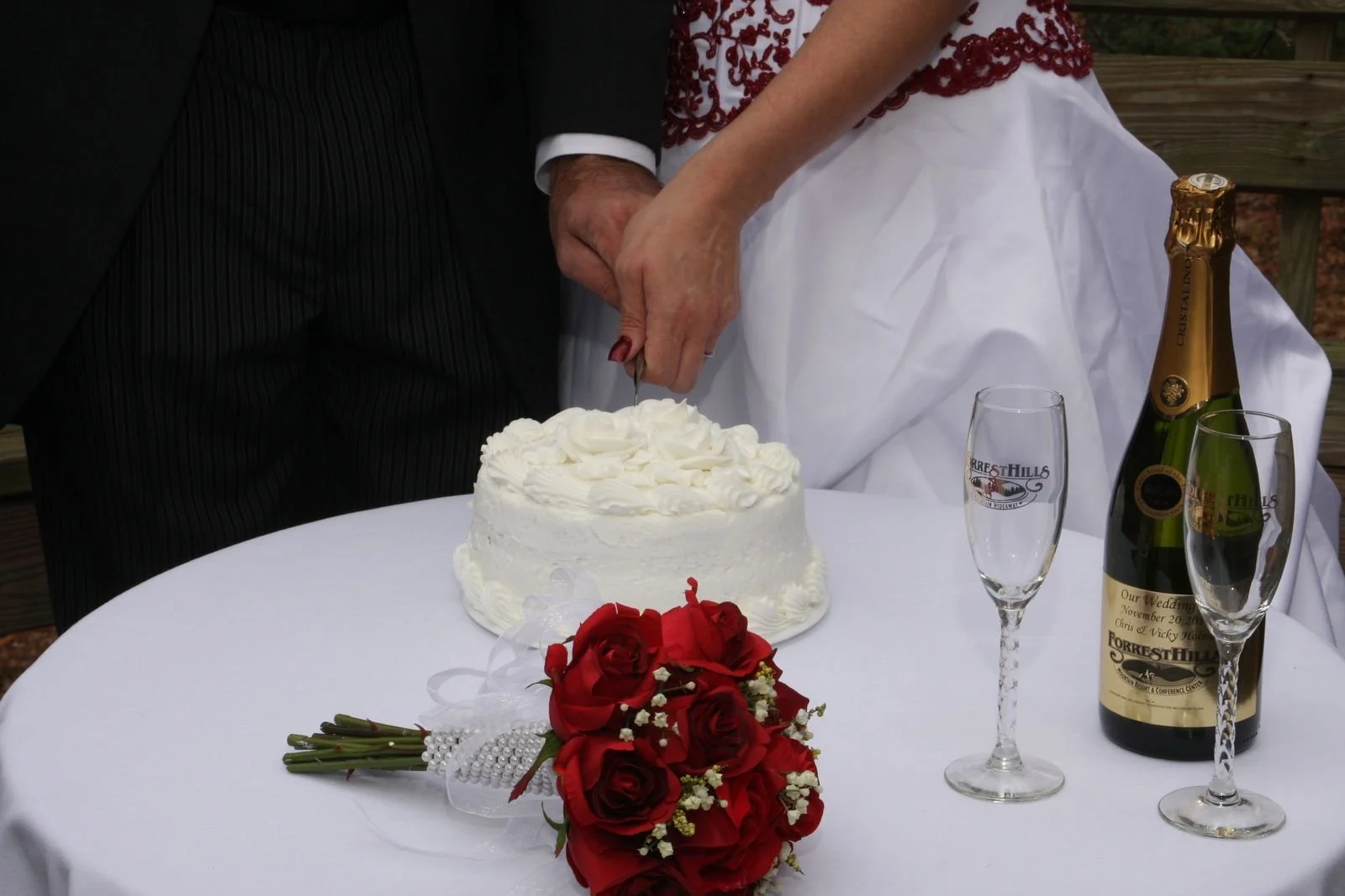 A bride and groom cut a wedding cake together at a wedding reception. The bride is wearing a white dress with red embroidery, and the groom is in a dark suit. There is a bouquet of red roses on the table, along with a bottle of champagne and two glas