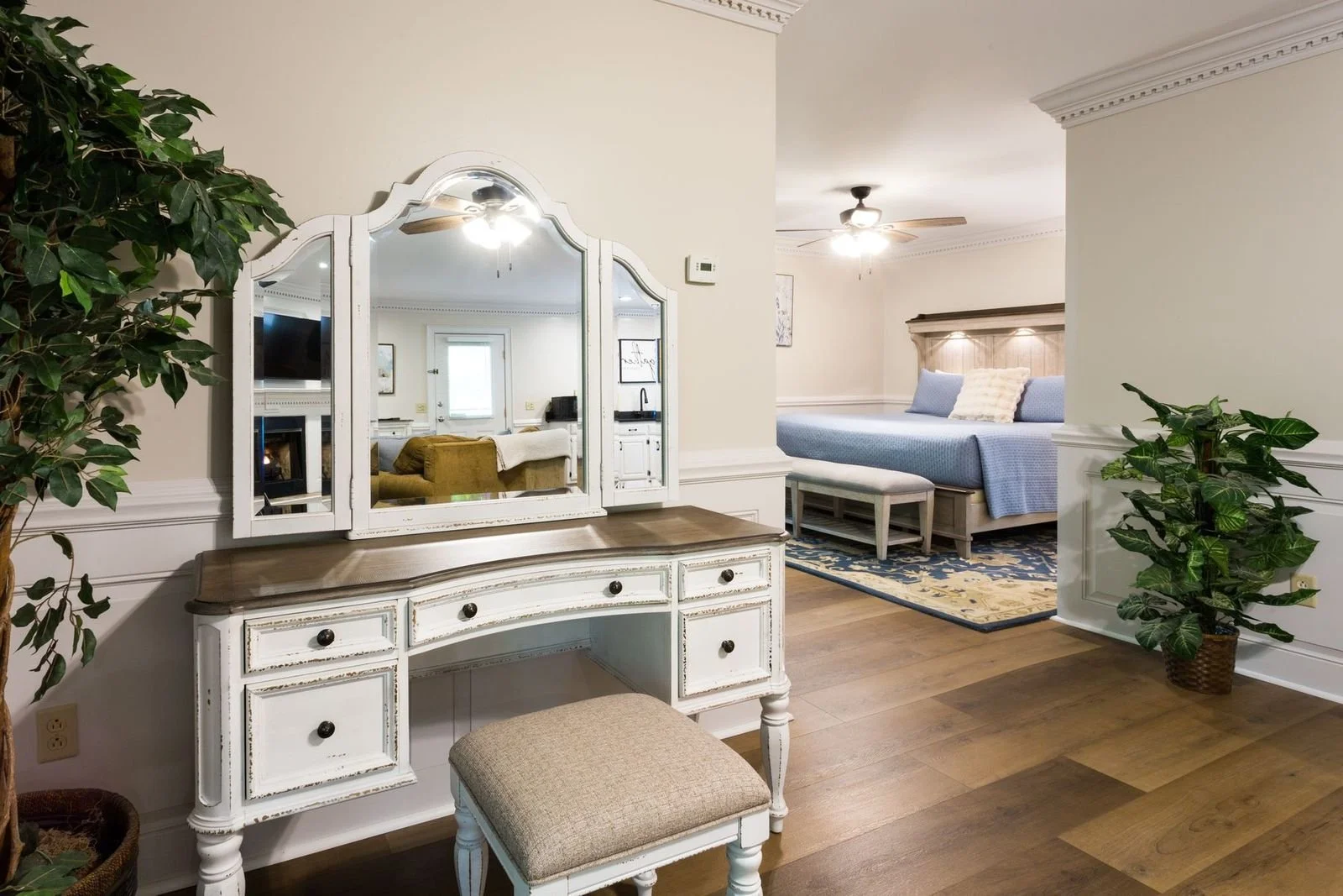 Interior view of a bedroom and dressing area showing a white vintage-style vanity with a large mirror, a beige cushioned stool, and a potted plant, with a bedroom in the background featuring a bed with blue bedding, a bench, ceiling fans, and framed artwork on the walls.