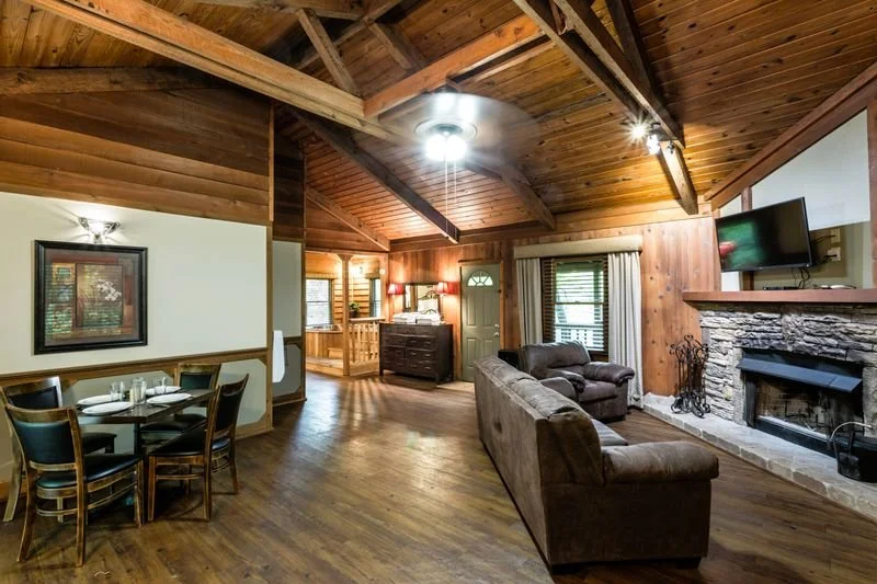 Living room with wooden vaulted ceiling, stone fireplace with mounted TV, two sofas, dining table with chairs, and hardwood floors.