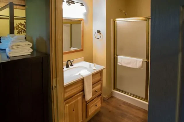 Bathroom with wooden vanity, mirror, towel ring, beige walls, glass shower with gold trim, and neatly folded towels on a dark cabinet.