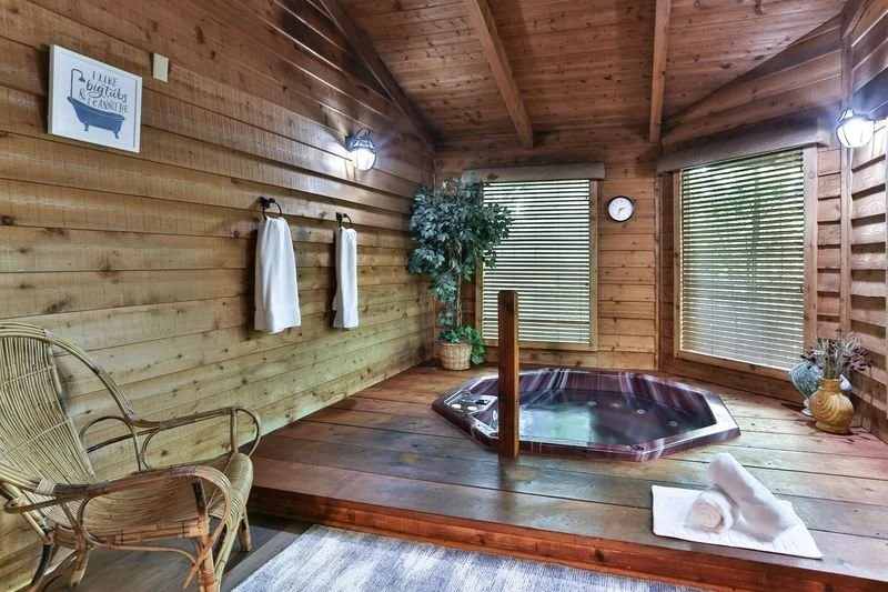 A cozy wooden room with a hot tub, wicker chair, potted plants, and towels.