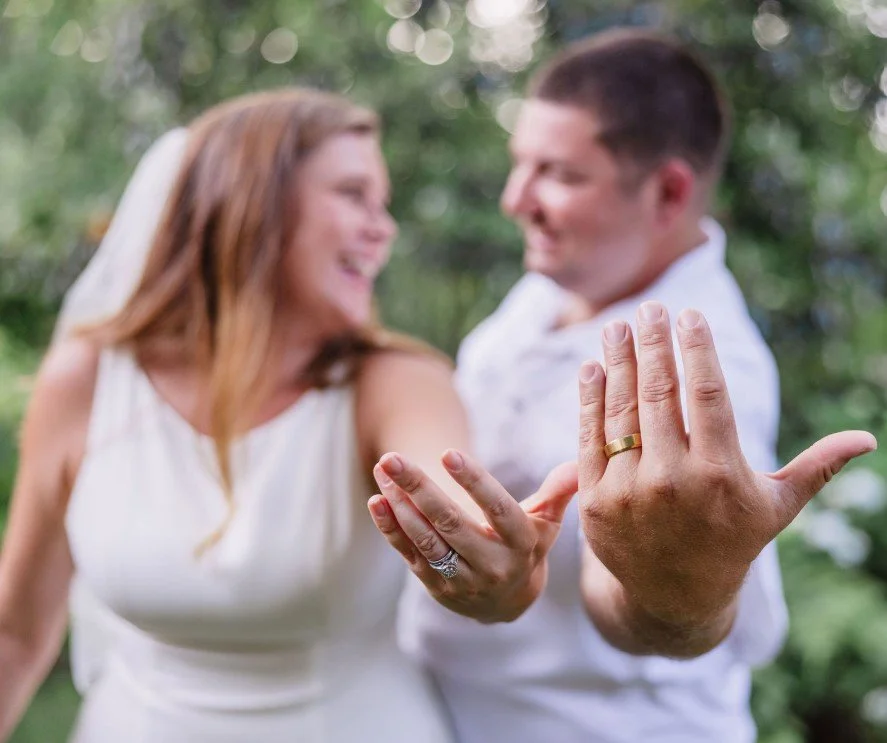 A happy couple shows off their wedding rings during an outdoor celebration with greenery in the background.
