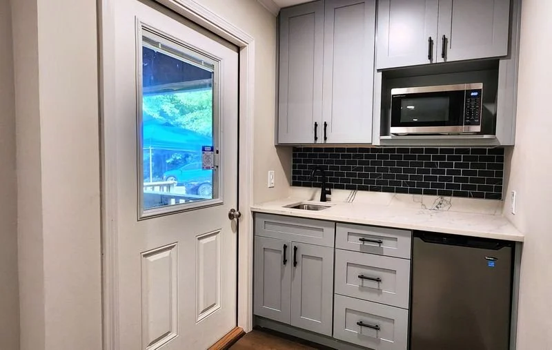 Small kitchen area with gray cabinets, black subway tile backsplash, microwave, mini fridge, and a door with a window.