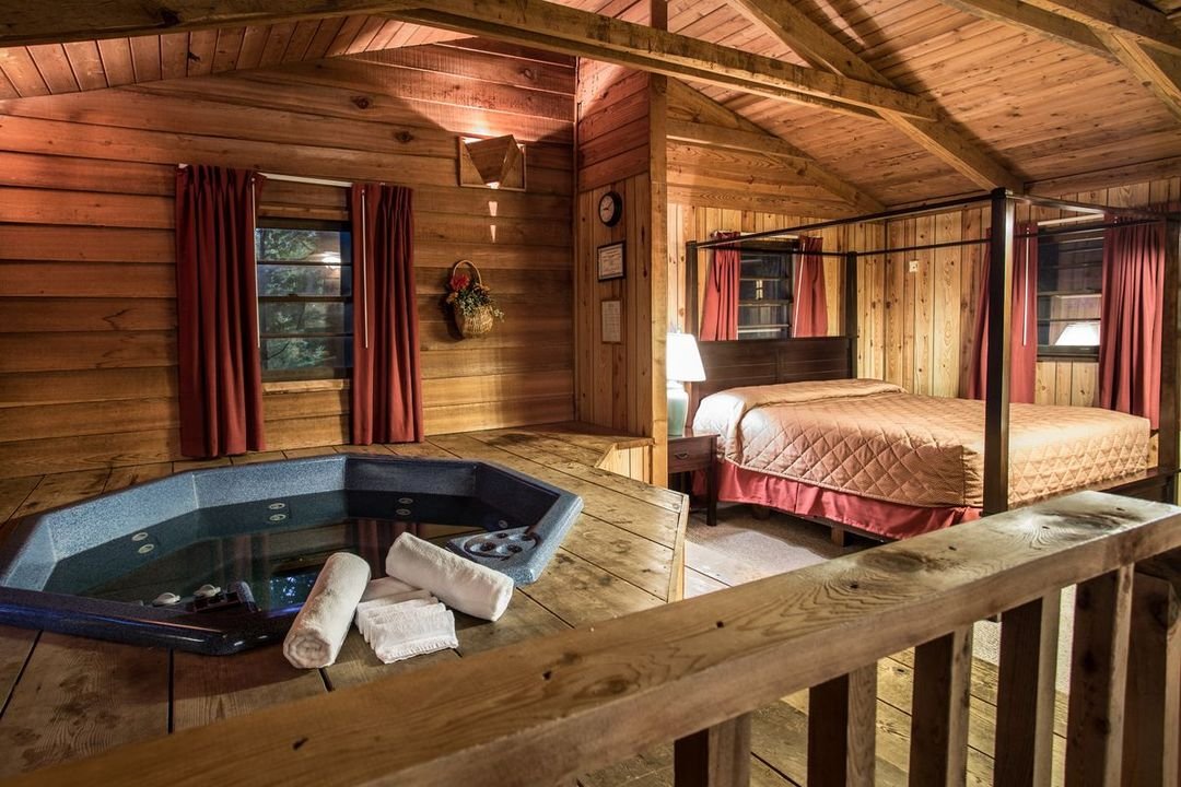 A cozy rustic bedroom with a hot tub, a large bed with a canopy frame, wooden walls, red curtains, and warm lighting.