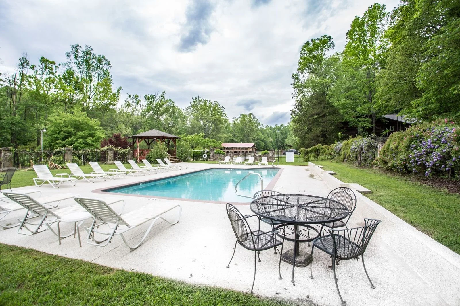 Outdoor swimming pool area with white lounge chairs, black metal table and chairs, surrounded by greenery and flowering bushes, with a gazebo and wooded area in the background.