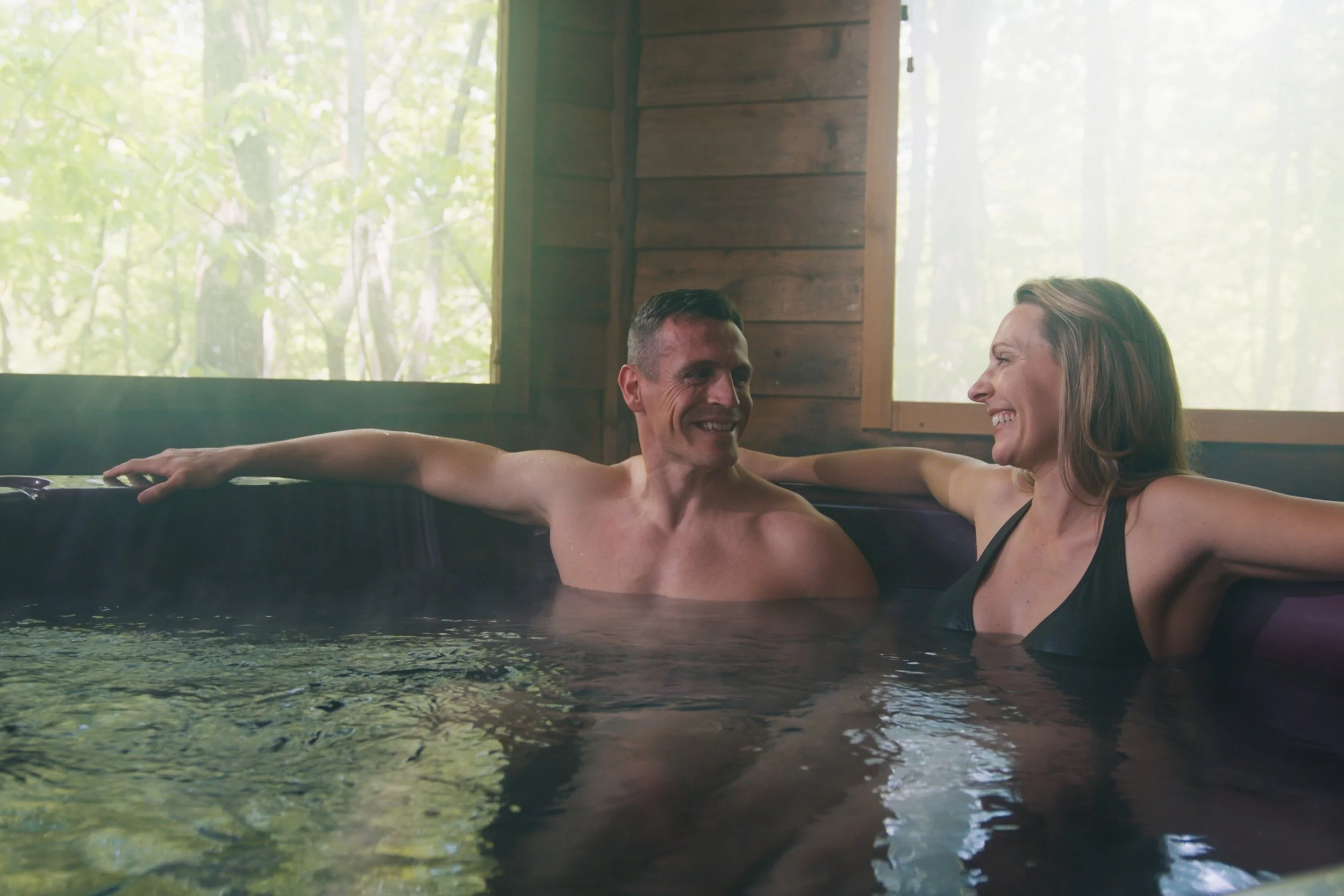 A man and woman relaxing together in a hot tub inside a wooden cabin with a window showing green trees outside.