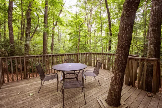 Wooden deck with three chairs and a table, surrounded by green trees in a forest.