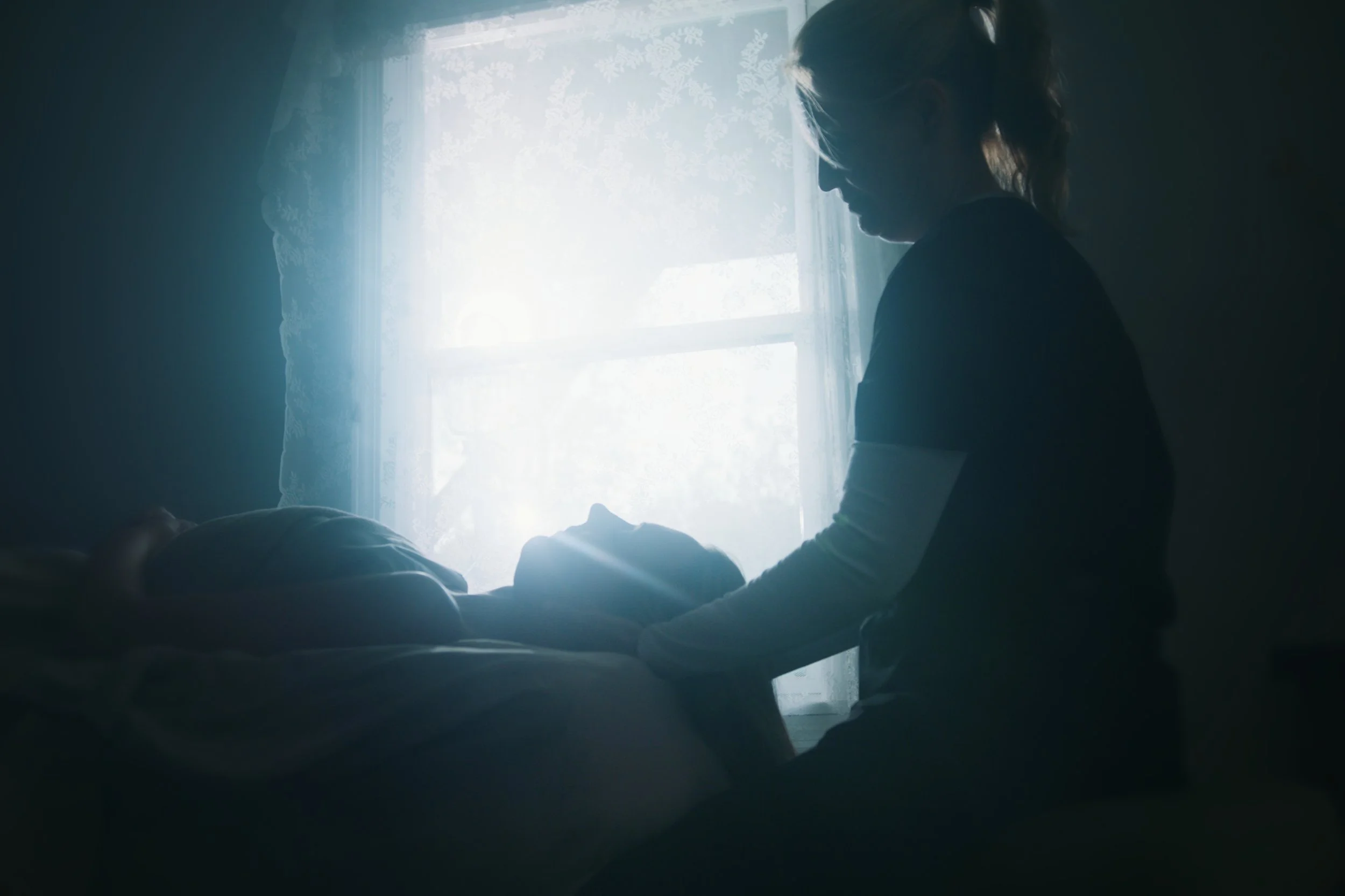 A woman looks down at a person lying on a bed, with sunlight coming through a window with lace curtains in the background.