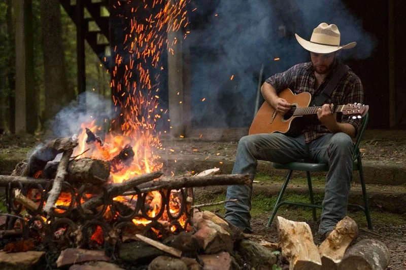 A man wearing a cowboy hat sitting on a green chair, playing an acoustic guitar near a campfire with logs and sparks, outdoors in a wooded area.