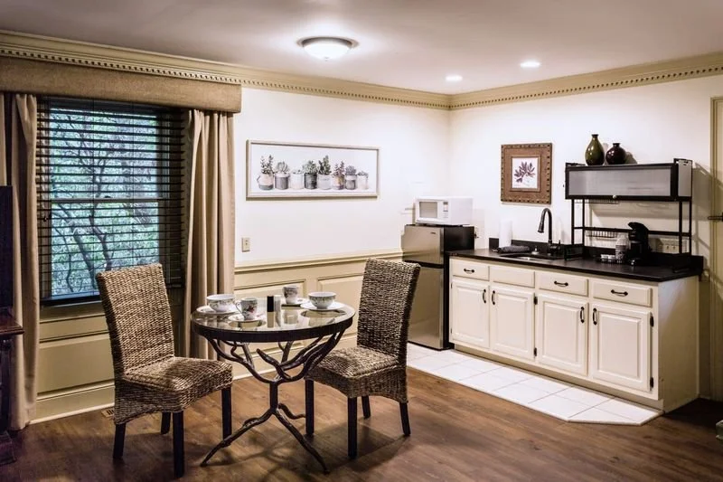 Cozy kitchen and dining area with a round table set for three, wicker chairs, a window with blinds, and white cabinets with black countertops under warm lighting.