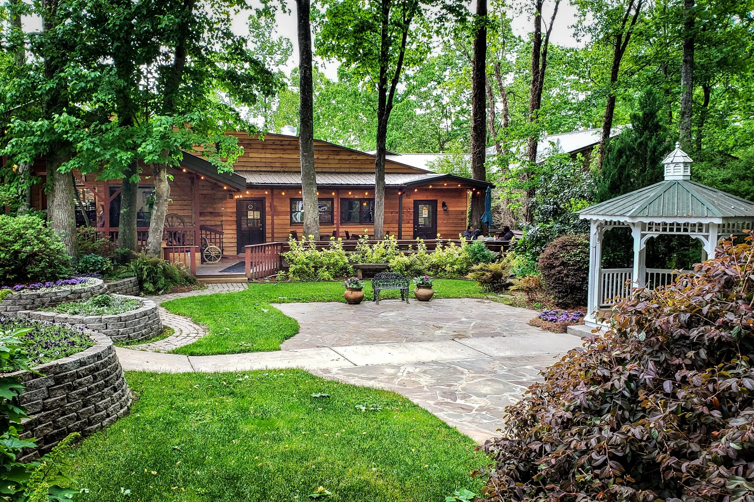 A lush backyard with a stone patio, surrounded by trees and colorful plants, featuring a wooden house with string lights and a white gazebo.