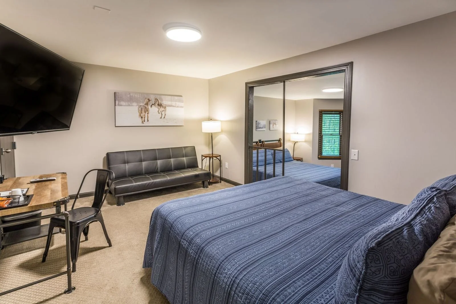 Interior of a bedroom with a large bed covered in a blue patterned bedspread, a black leather futon, a wooden desk with a black chair, a flat-screen TV, nightstand with lamp, a large mirror, and a window with blinds.