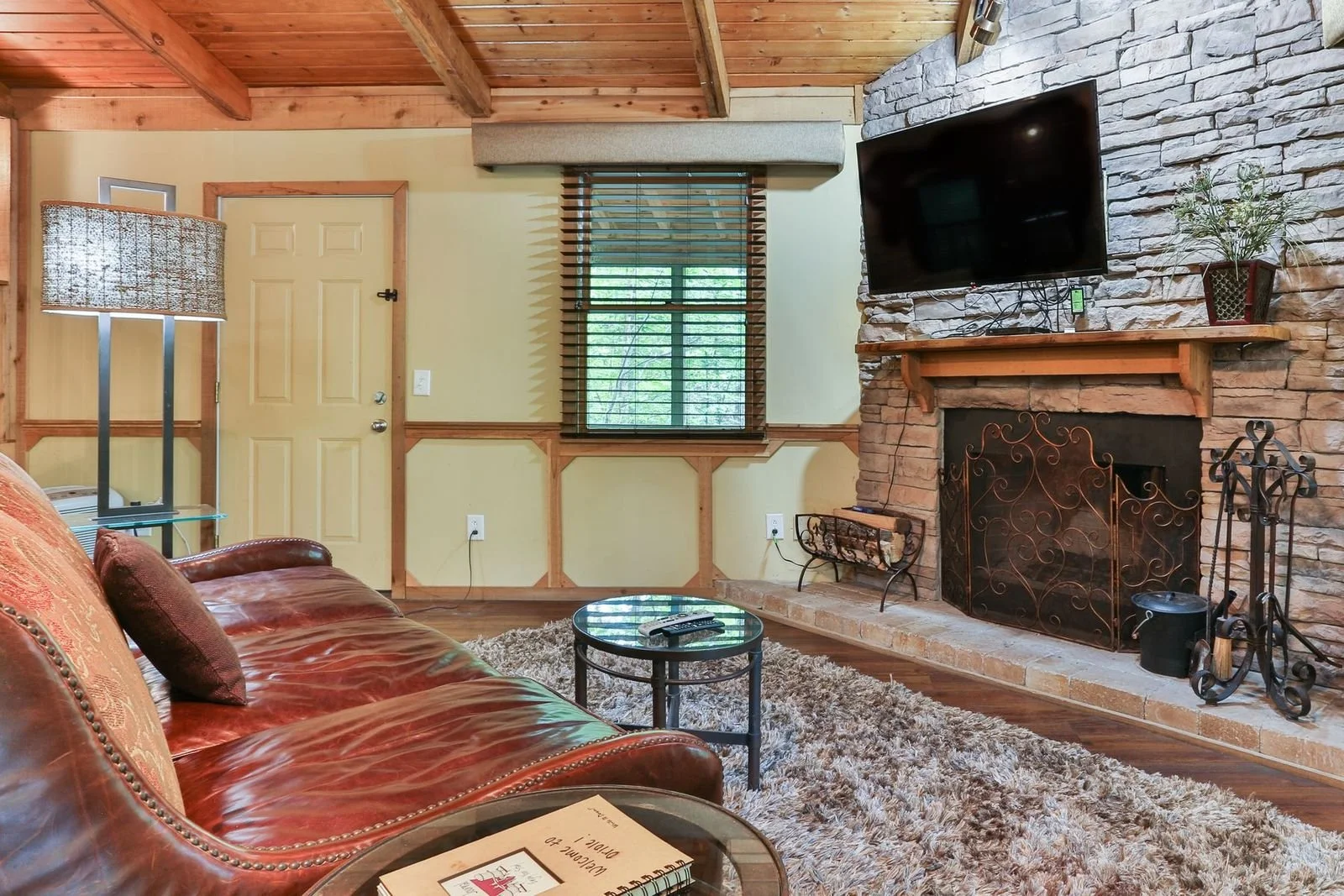 Living room with leather sofa, glass coffee table, wood and stone fireplace, wall-mounted TV, window with wooden blinds, wooden ceiling beams, and a shaggy area rug.