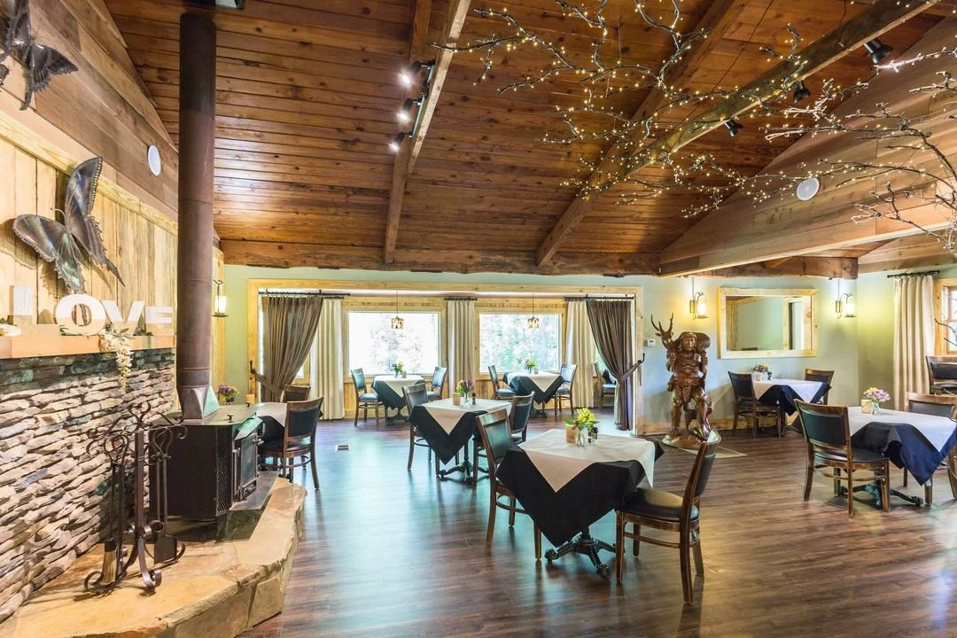 Dining room with wooden ceiling, string lights, and several tables with black and white tablecloths, decorated with small flower arrangements, with curtains and windows in the background.