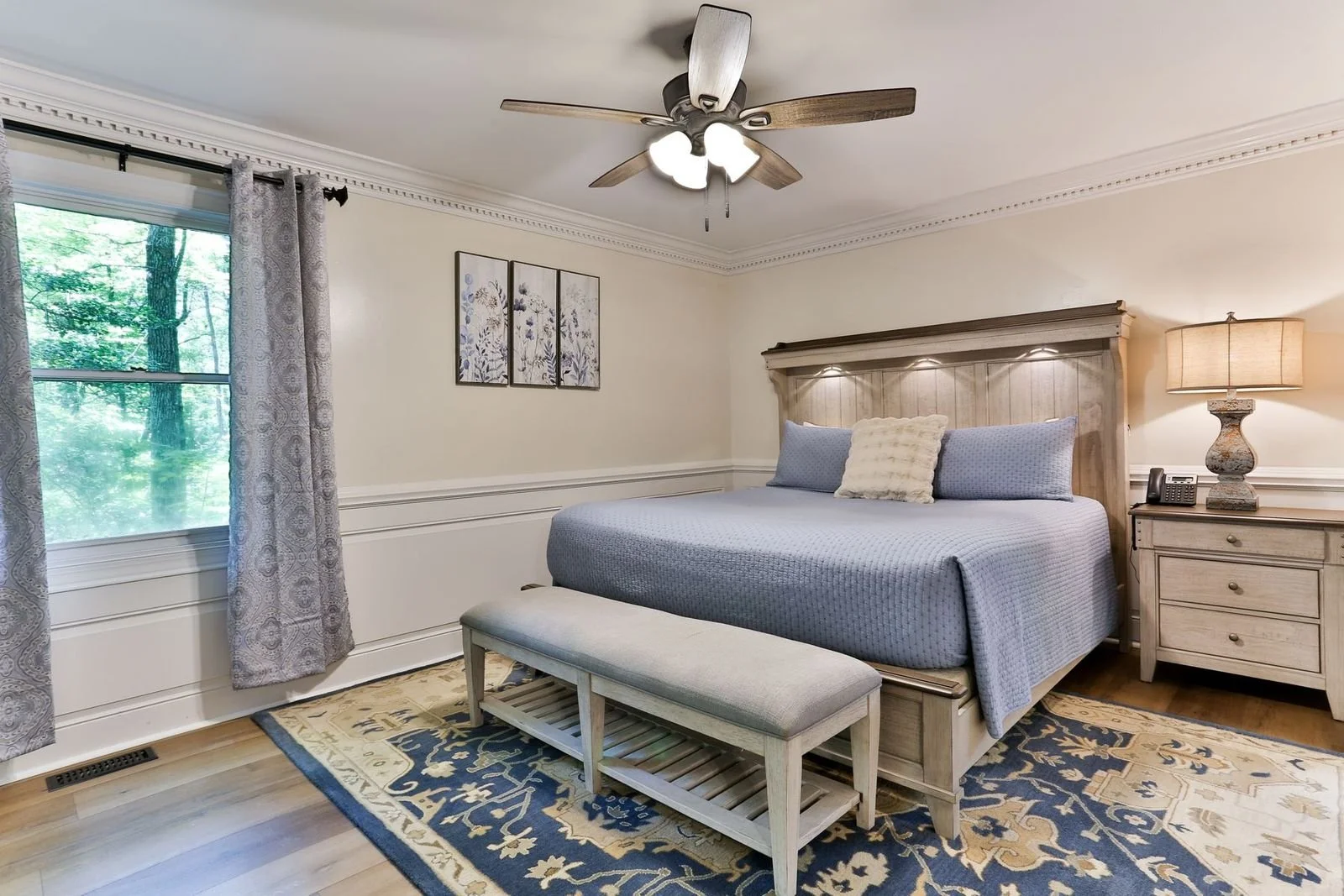 Bedroom with a large bed, wooden headboard with built-in lights, blue bedding, and a cream decorative pillow. A window with patterned curtains showing trees outside, a nightstand with a lamp and phone, a framed wall art, and a ceiling fan with lights