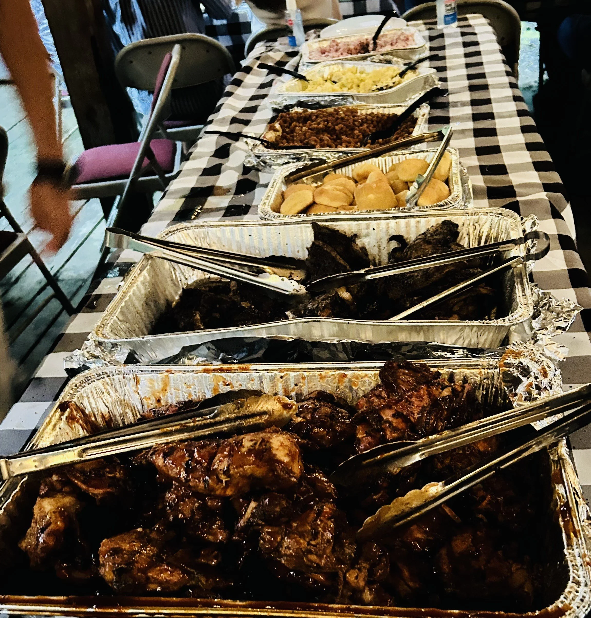 Buffet table with various dishes in aluminum trays at a gathering, checkered tablecloth, and chairs in the background.