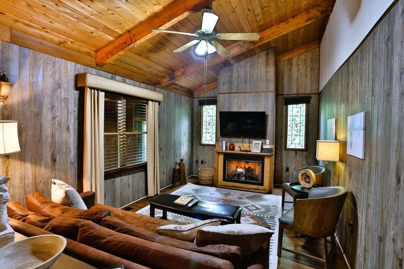 Cozy living room with wood-paneled walls and ceiling, a brown sectional sofa, a coffee table, a fireplace, and a flat-screen TV, lit by lamps and natural light through windows.