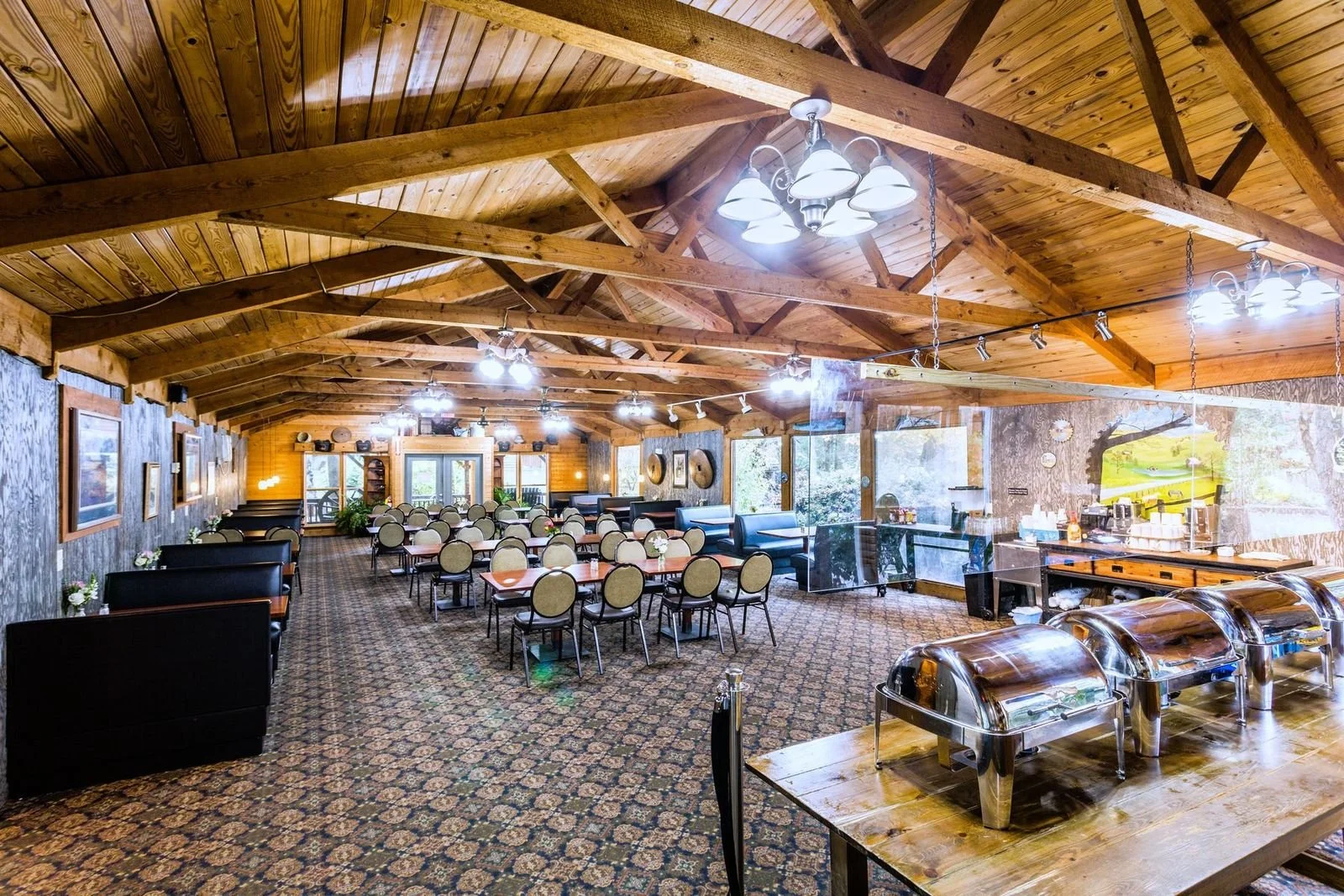 Spacious dining area with wooden ceilings, multiple hanging lights, and round tables with chairs. There are large windows allowing natural light, and a self-serve food station with chafing dishes.