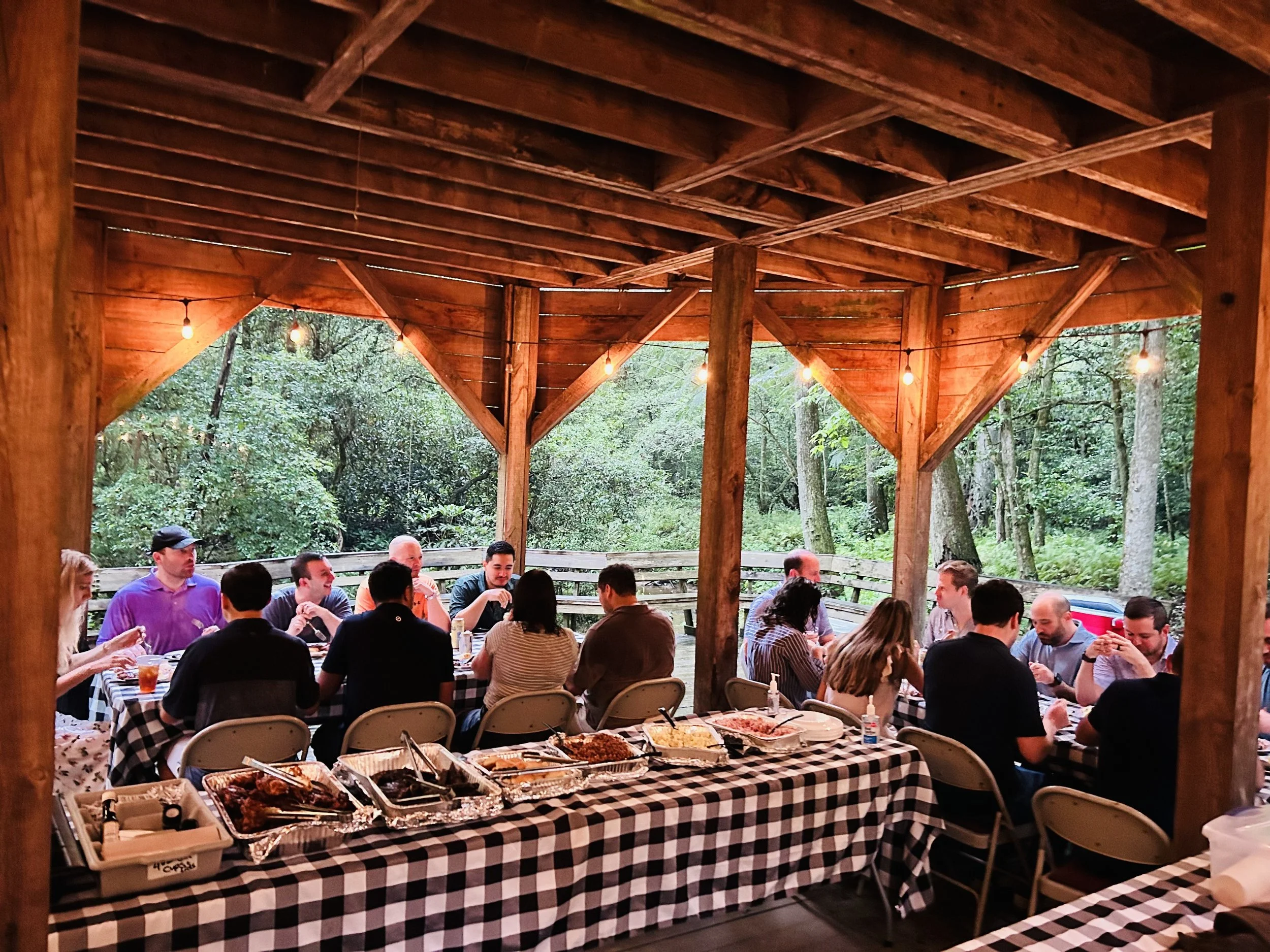Gathering of people sitting under a wooden pavilion with string lights, enjoying a meal with various dishes on a table covered with a black and white checkered tablecloth, surrounded by a forested outdoor setting.
