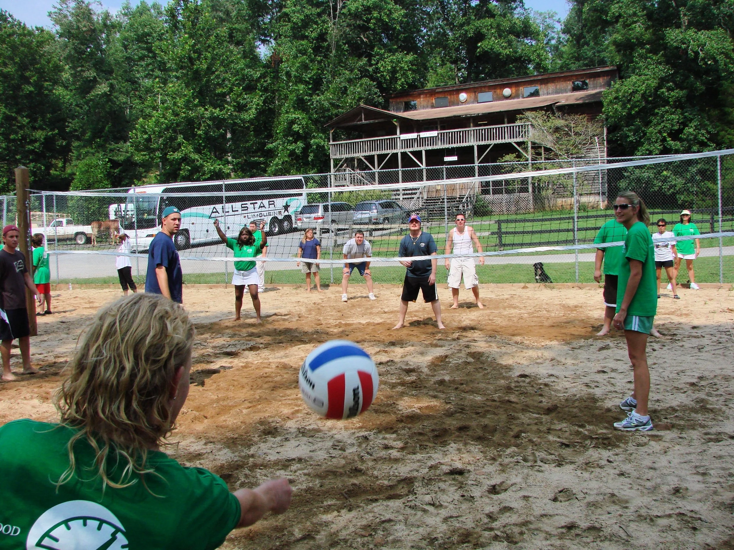 People playing volleyball on a sandy court outdoors, surrounded by a chain-link fence with lush green trees in the background.